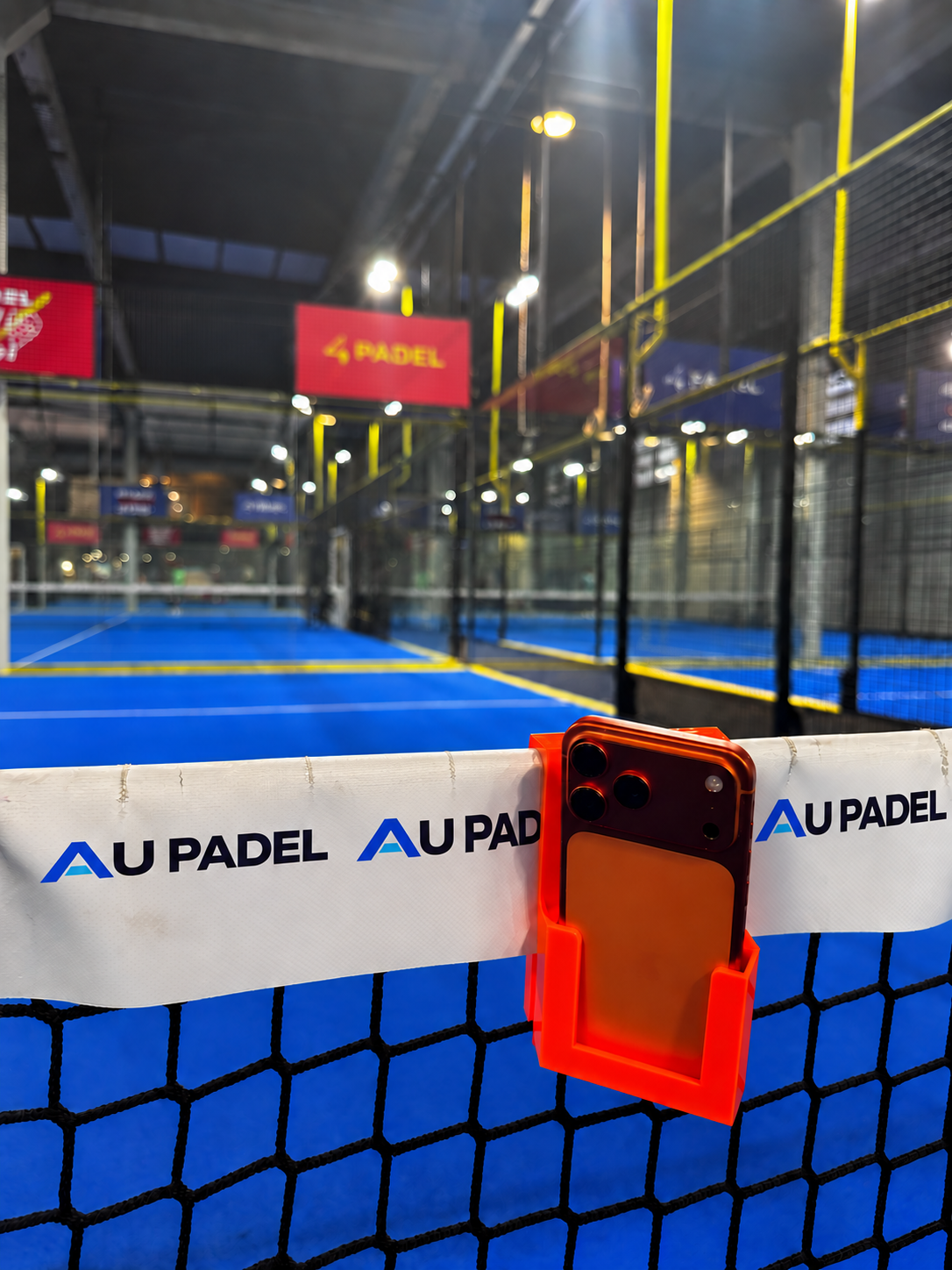 Smartphone posé verticalement dans le support PAD’VIEWS orange sur un filet AU PADEL — photo retouchée.